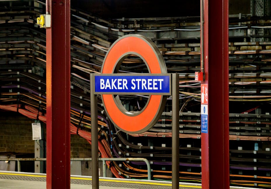 A dimly lit underground subway platform at Baker Street station with a vaulted brick ceiling and arched glass skylights allowing some natural light. Several illuminated signs display the station name